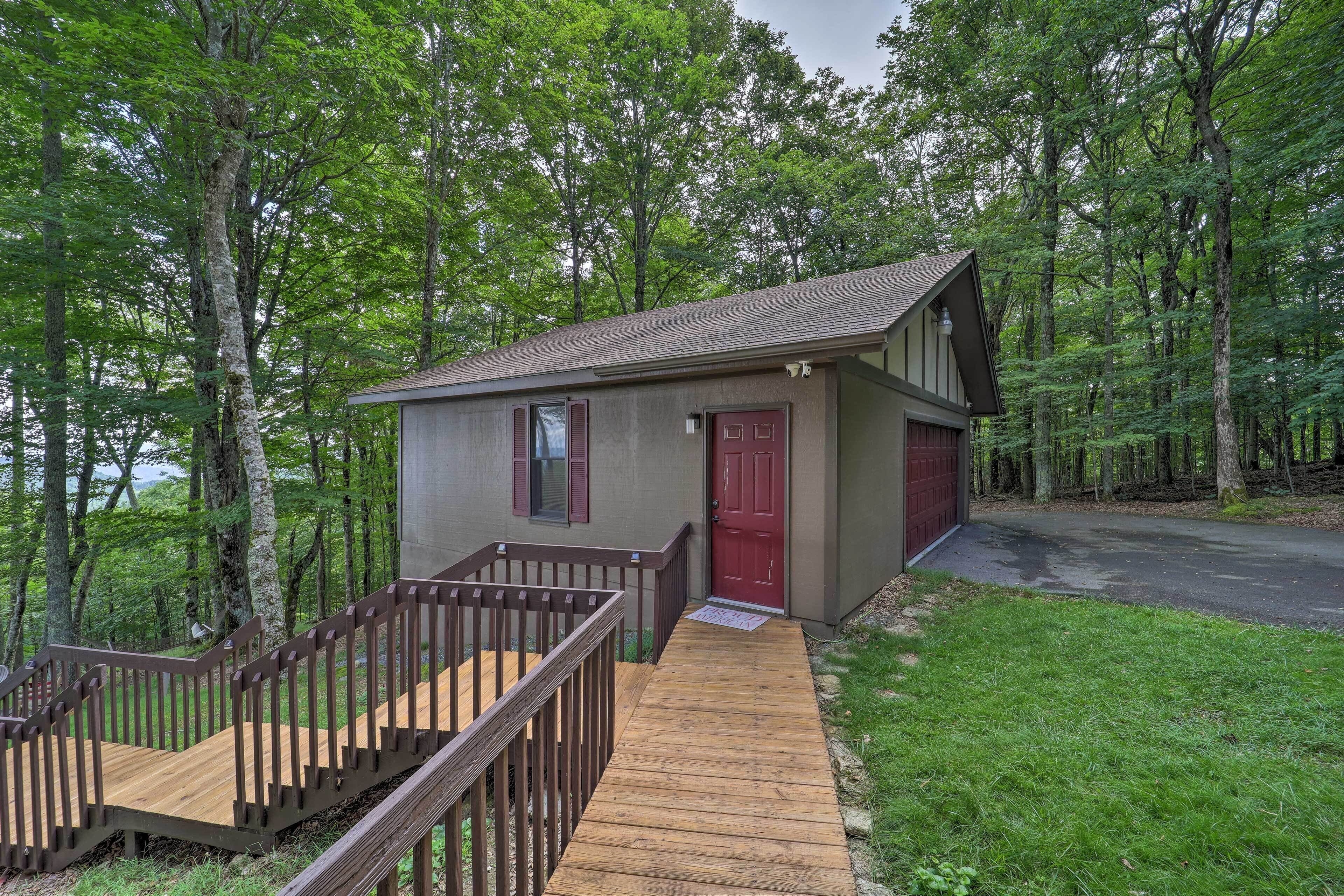 A small cabin with a wooden deck and a red door, surrounded by a forested area.