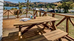Una terraza de madera con una mesa de picnic, rodeada por una barandilla de madera, con vista a un lago donde se observan vehículos recreativos estacionados.