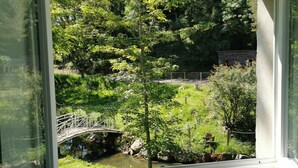 Une vue depuis une fenêtre à cadre blanc, donnant sur un paysage verdoyant et luxuriant, avec des arbres, un petit pont et un étang.
