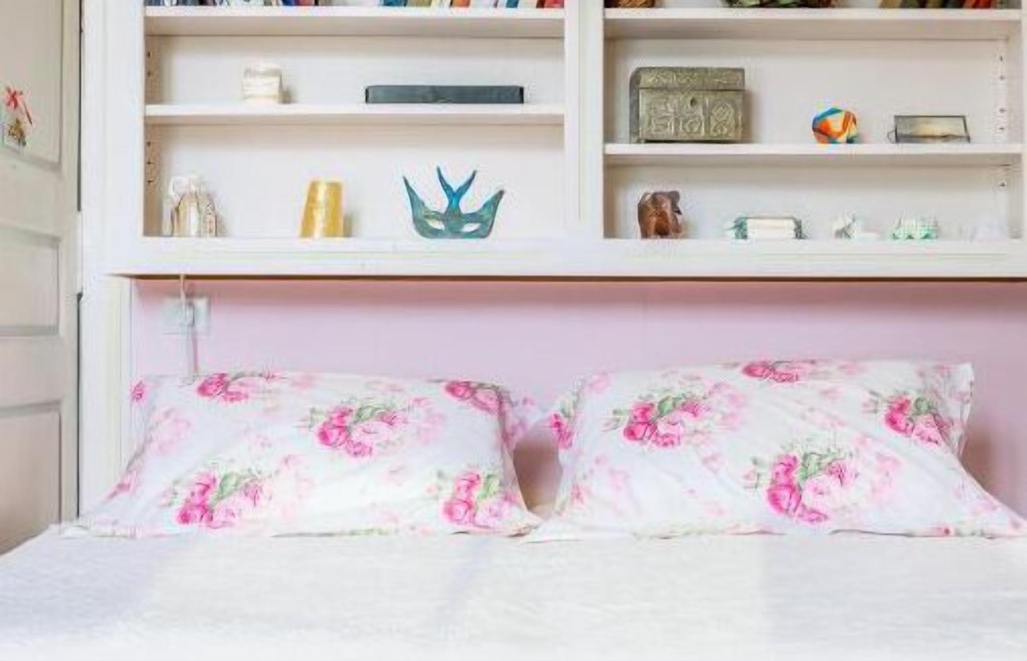 Een slaapkamer met een bed, bloemenkussens, een plank met diverse spullen en een roze hoofdeinde.