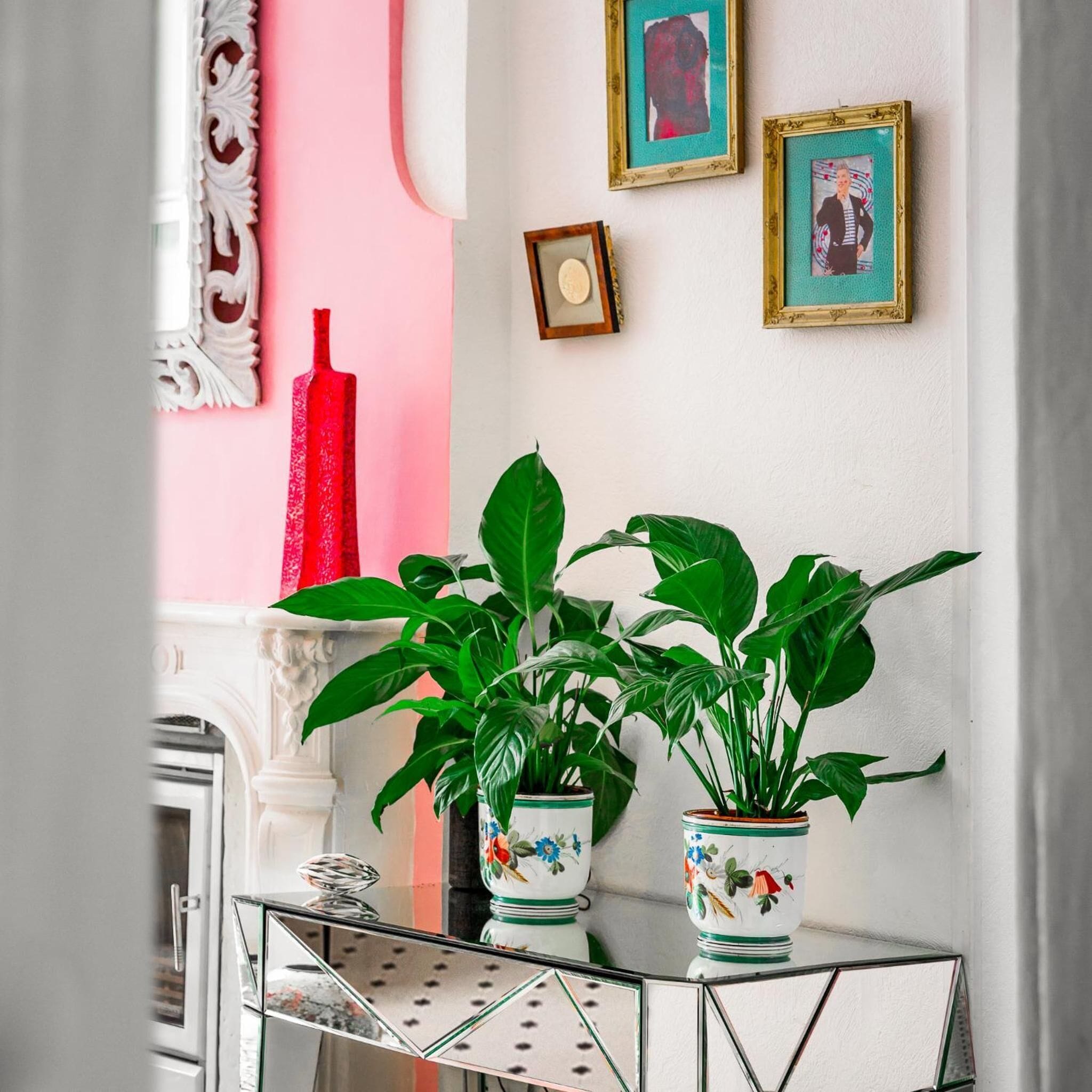 A room with a white fireplace, a pink wall, and a white column with ornate detailing. There are three framed pictures on the wall and a red vase on the mantel.