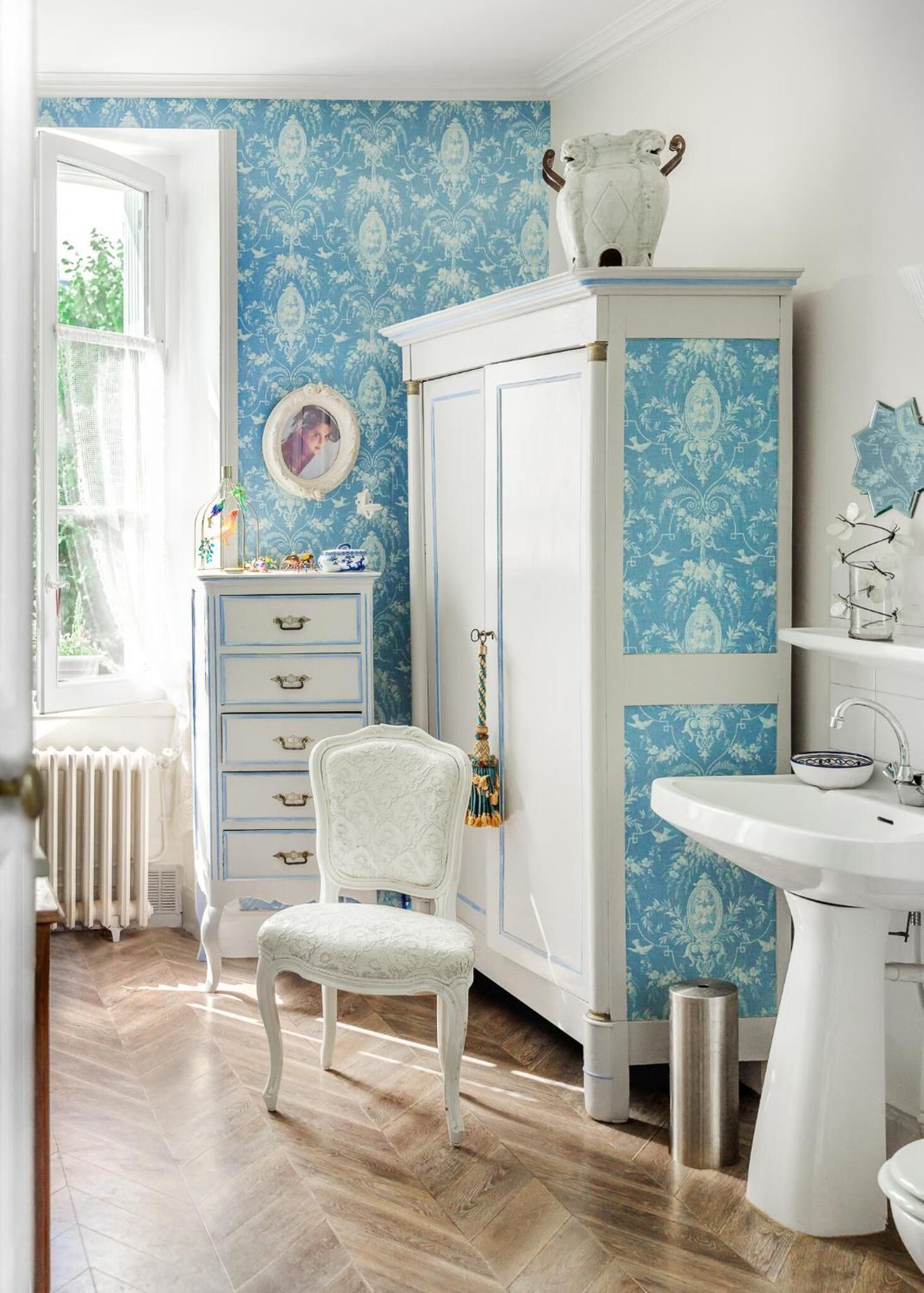 A room with a blue patterned wallpaper, a white cabinet with a decorative top, a white chair, and a white sink.