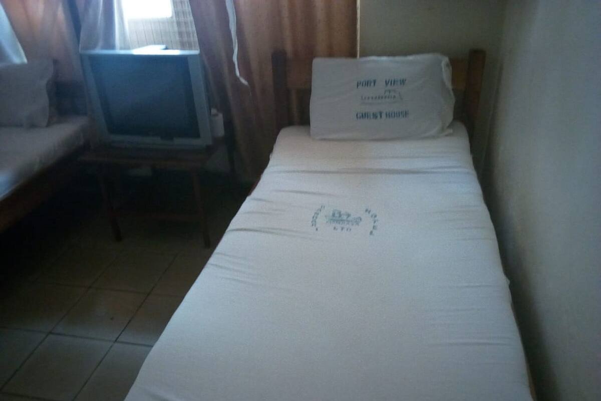 Ein Einzelbett mit einem Kopfkissen, auf dem "Port View Guest House" steht, und ein kleiner Tisch mit einem Fernseher.