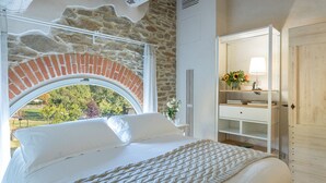 Quarto com janela em arco de tijolos, cama com roupa de cama branca, guarda-roupa de madeira e vista para o verde lá fora.