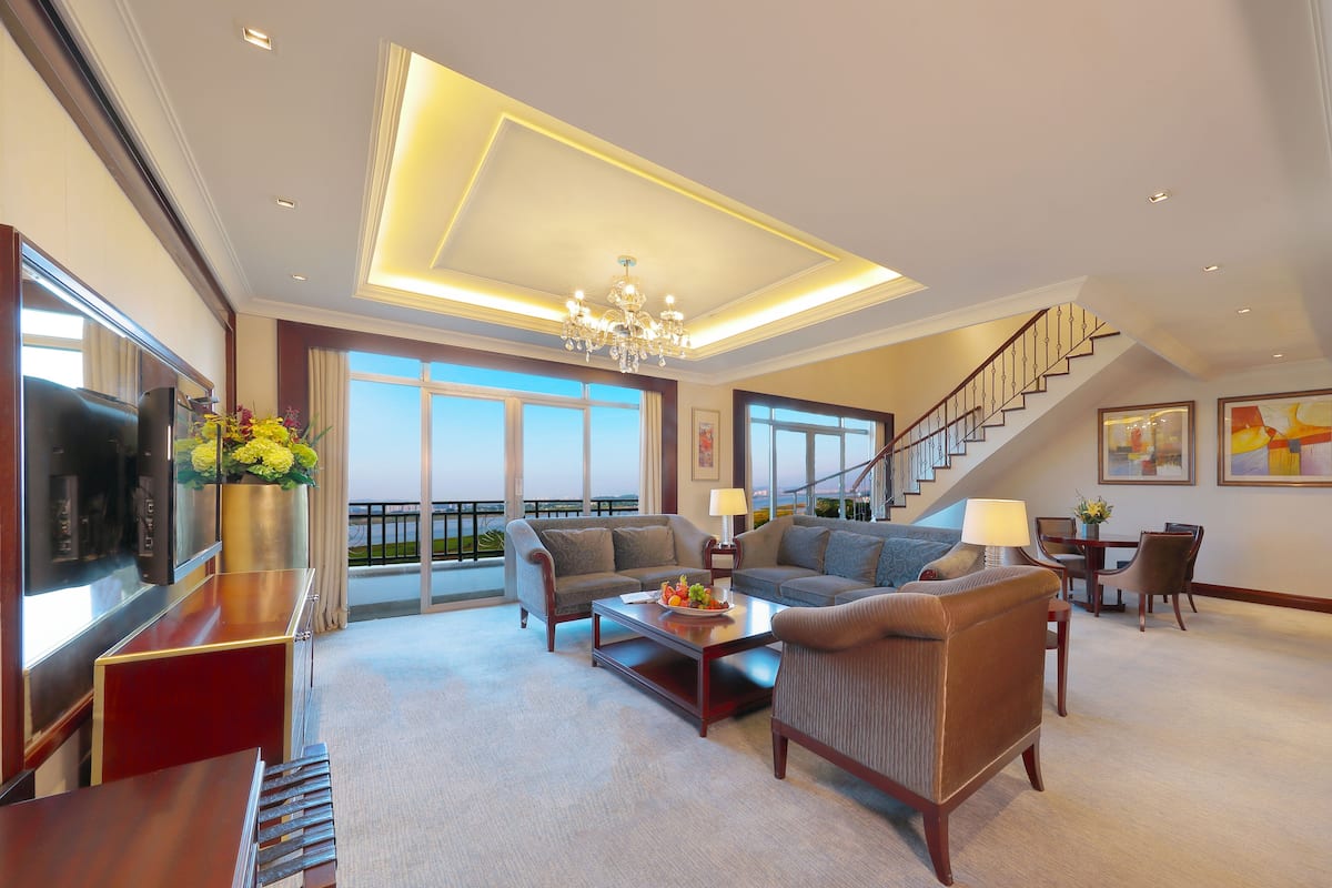 A spacious living room with a large sofa set, a coffee table, and a TV. There is a staircase leading to an upper level with a balcony.