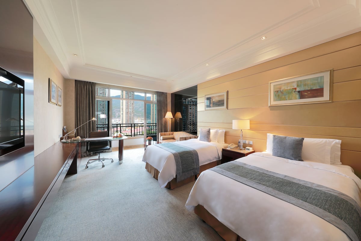 A hotel room with two beds, a desk, a television, and a large window with a view of buildings.