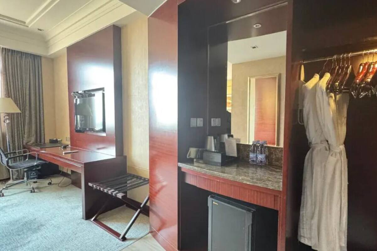 A hotel room with a desk, chair, and a mirror. There is a coat rack with clothes and a small refrigerator.