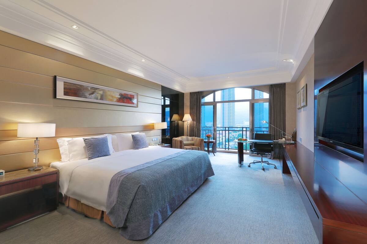 A hotel room with a large bed, a desk with a chair, a TV, and a view of the city.