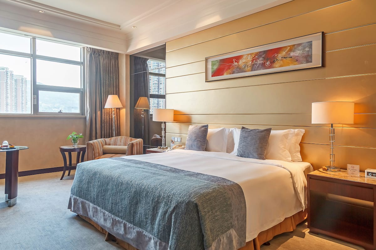A hotel room with a large bed, two bedside lamps, a nightstand, a chair, a small table with a plant, and a painting on the wall.