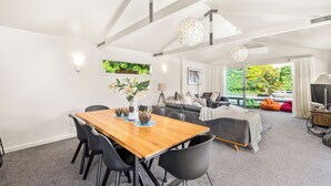 A modern living room with a wooden dining table, black chairs, a grey sofa, and a large window with a view of greenery.