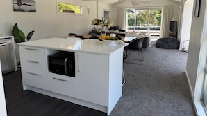 A modern kitchen with white cabinetry, a built-in oven, and a large island. There is a dining area with a wooden table and chairs, and a view of the outdoors through large windows.