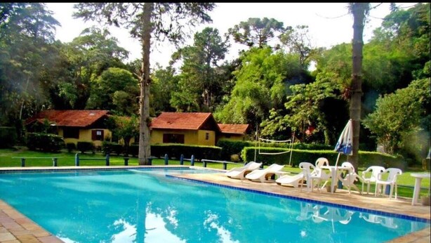 A swimming pool with lounge chairs and a small table, surrounded by greenery and a few buildings. - Pousada Sítio da Ponte (Itatiaia)
