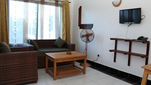 A living room with a sofa, a wooden table, a standing fan, a TV mounted on the wall, and a window with curtains. - Rock Galana Holiday Apartments (Diani Beach)