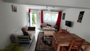 A living room with a sofa, a dining table with chairs, a TV, and a plant. - Rock Galana Holiday Apartments (Diani Beach)