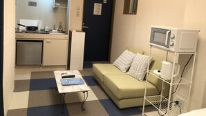 A compact living space with a kitchenette, a sofa with patterned cushions, a small table with a remote control, and a microwave on a stand. - Oval Tokyo Ikesan 201 (Tokyo)