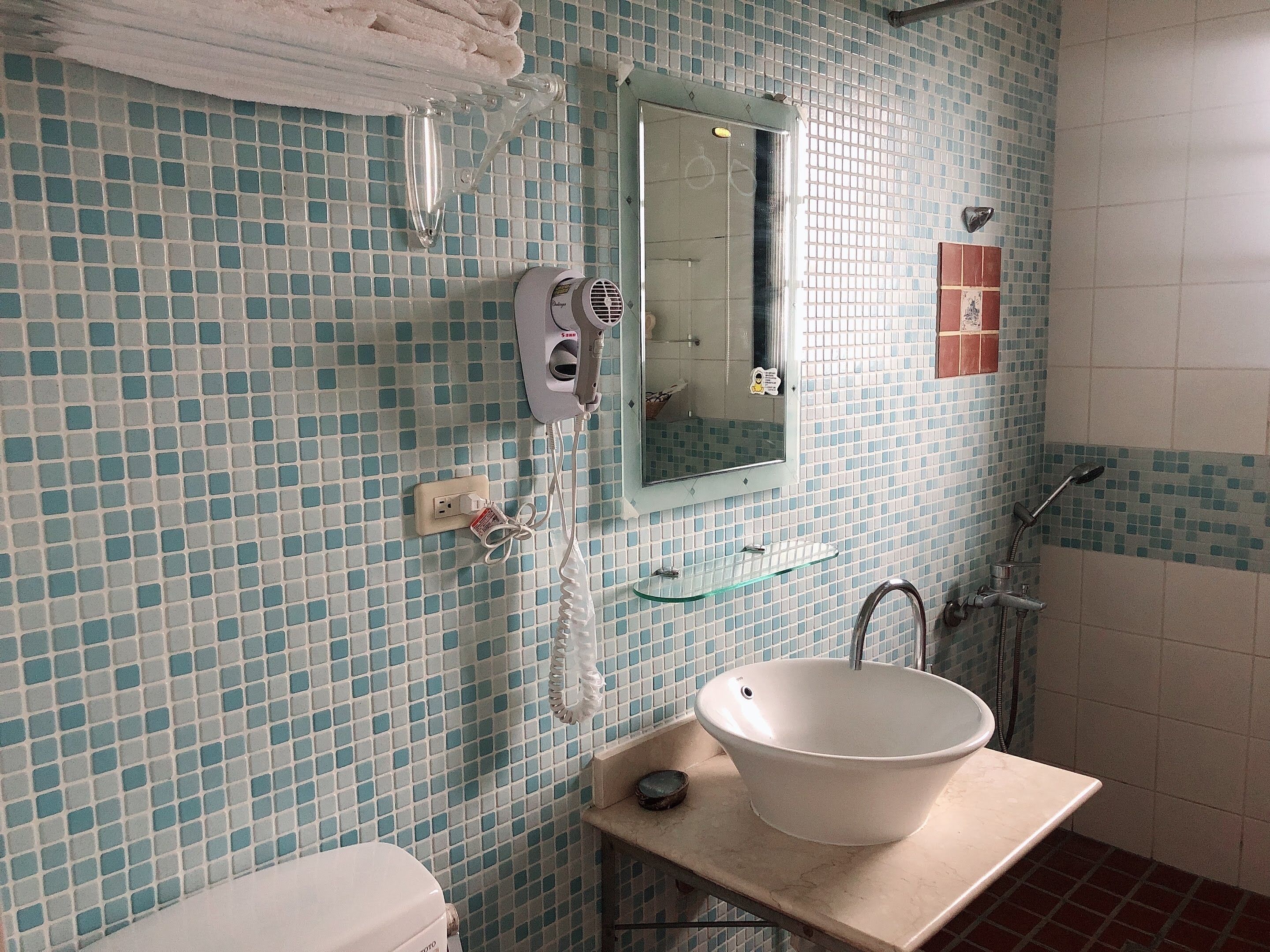 A bathroom with a tiled wall, a sink with a basin, a mirror, a towel rack, and a shower area.