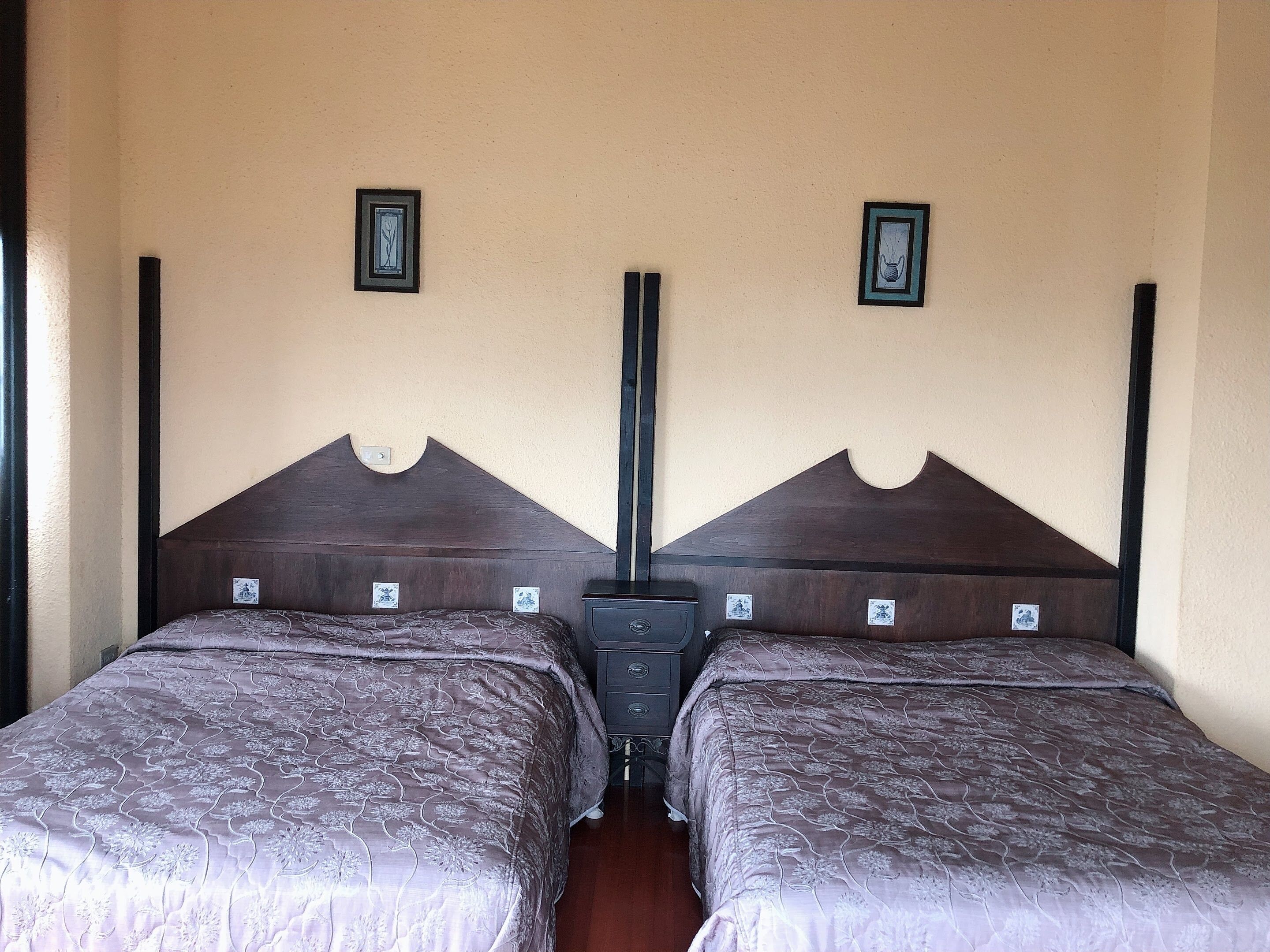 A double room with two beds, each with a wooden headboard and a floral-patterned bedspread. There are two small framed pictures on the wall above each bed.