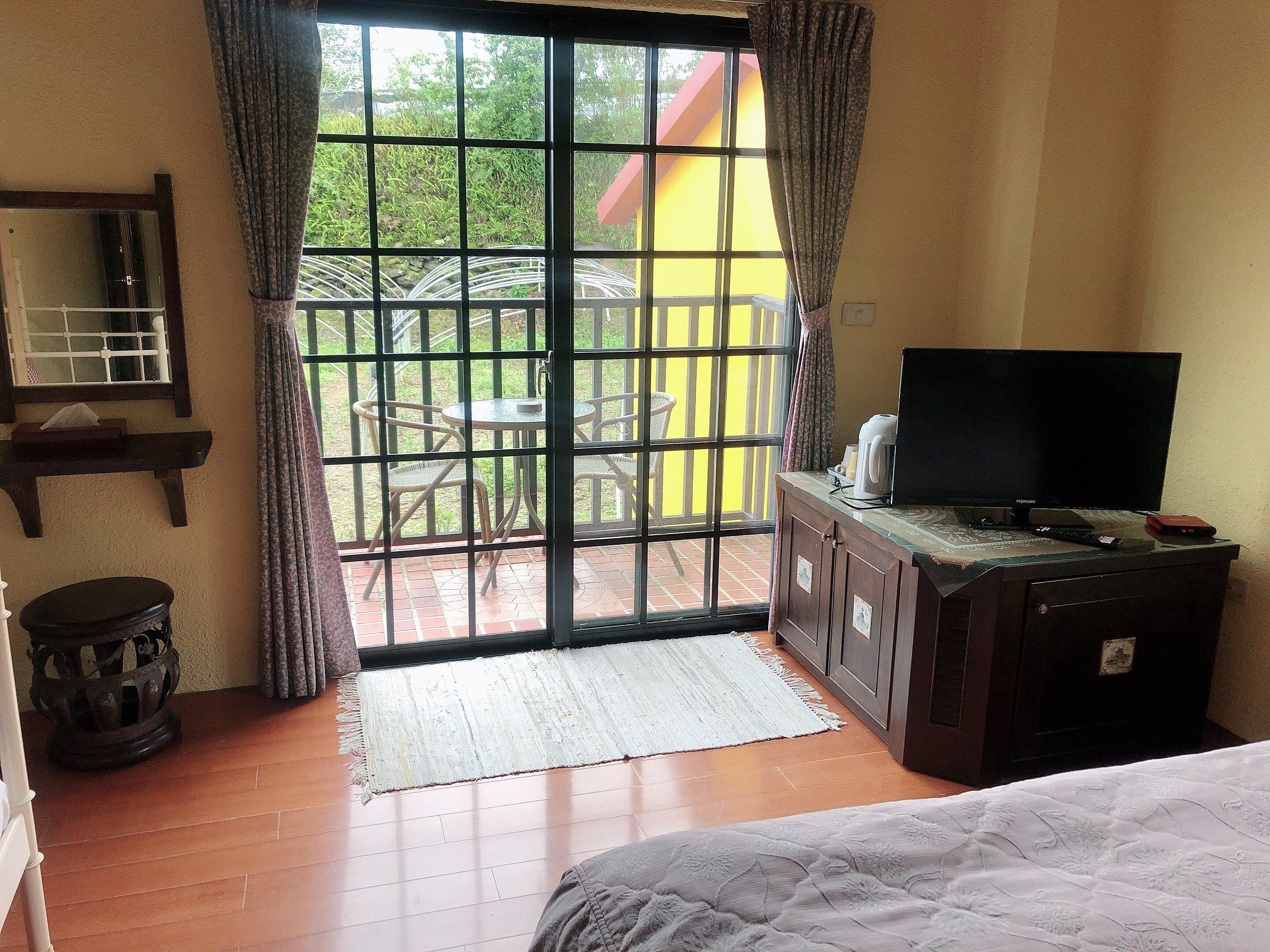 A room with a large sliding glass door leading to an outdoor area with a table and chairs. There is a bed with a white comforter, a television on a wooden cabinet, and a small round table with a lamp.