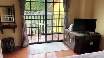A room with a large sliding glass door leading to an outdoor area with a table and chairs. There is a bed with a white comforter, a television on a wooden cabinet, and a small round table with a lamp.