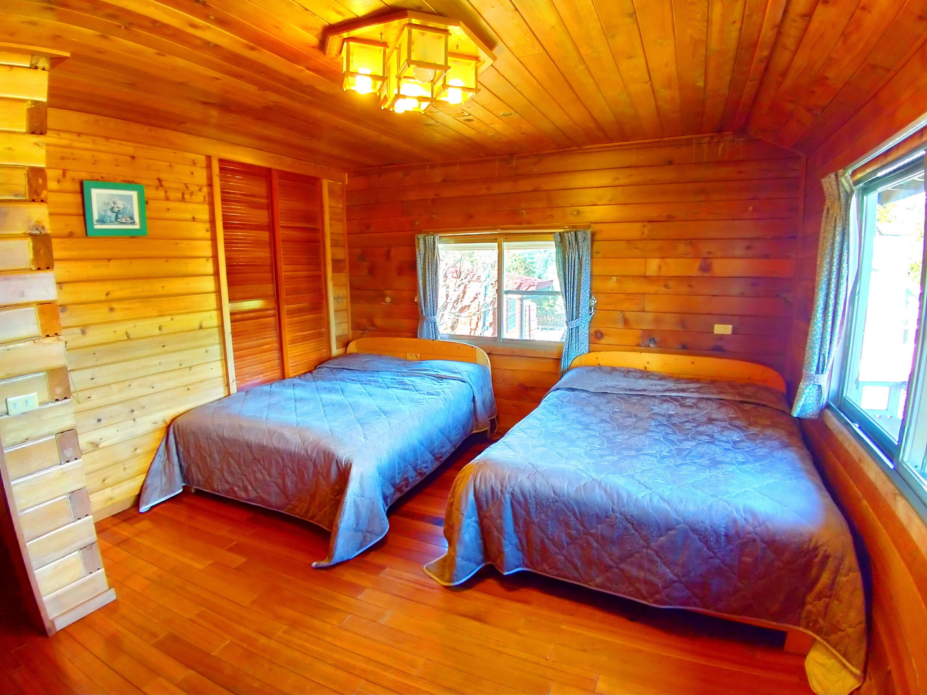 A cabin with two beds, wooden walls, a window with curtains, and a wooden ceiling with a light fixture.