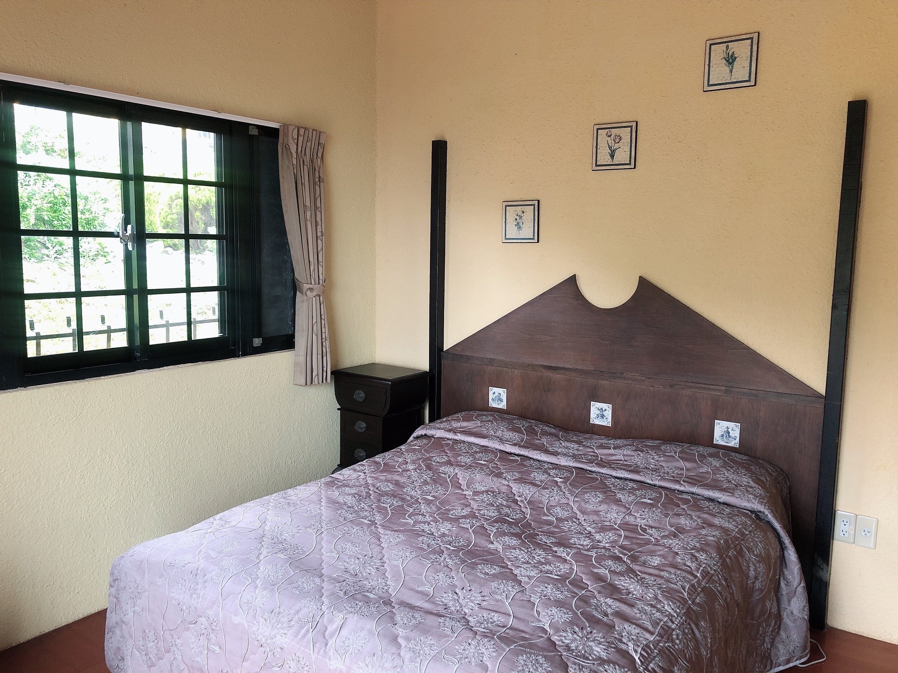 A bedroom with a bed, a nightstand, a window with curtains, and three wall-mounted pictures.