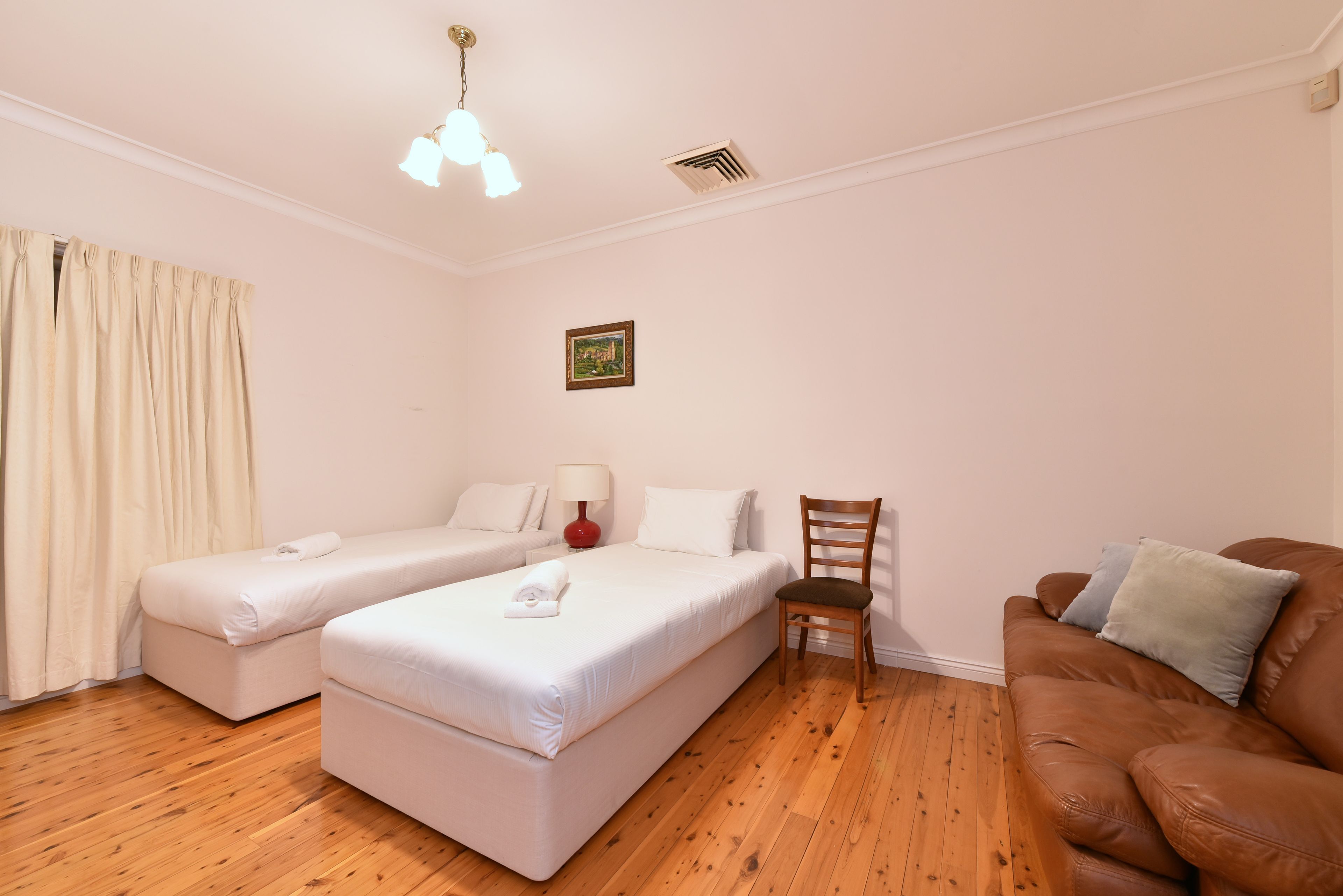 A hotel room with two single beds, a wooden floor, a red lamp, a framed picture on the wall, and a brown leather sofa.
