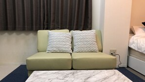 Ein kleines, modernes Hotelzimmer mit einem grünen Sofa, zwei gemusterten Kissen, einem Marmor-Couchtisch und einem Bett mit weißer Bettwäsche.