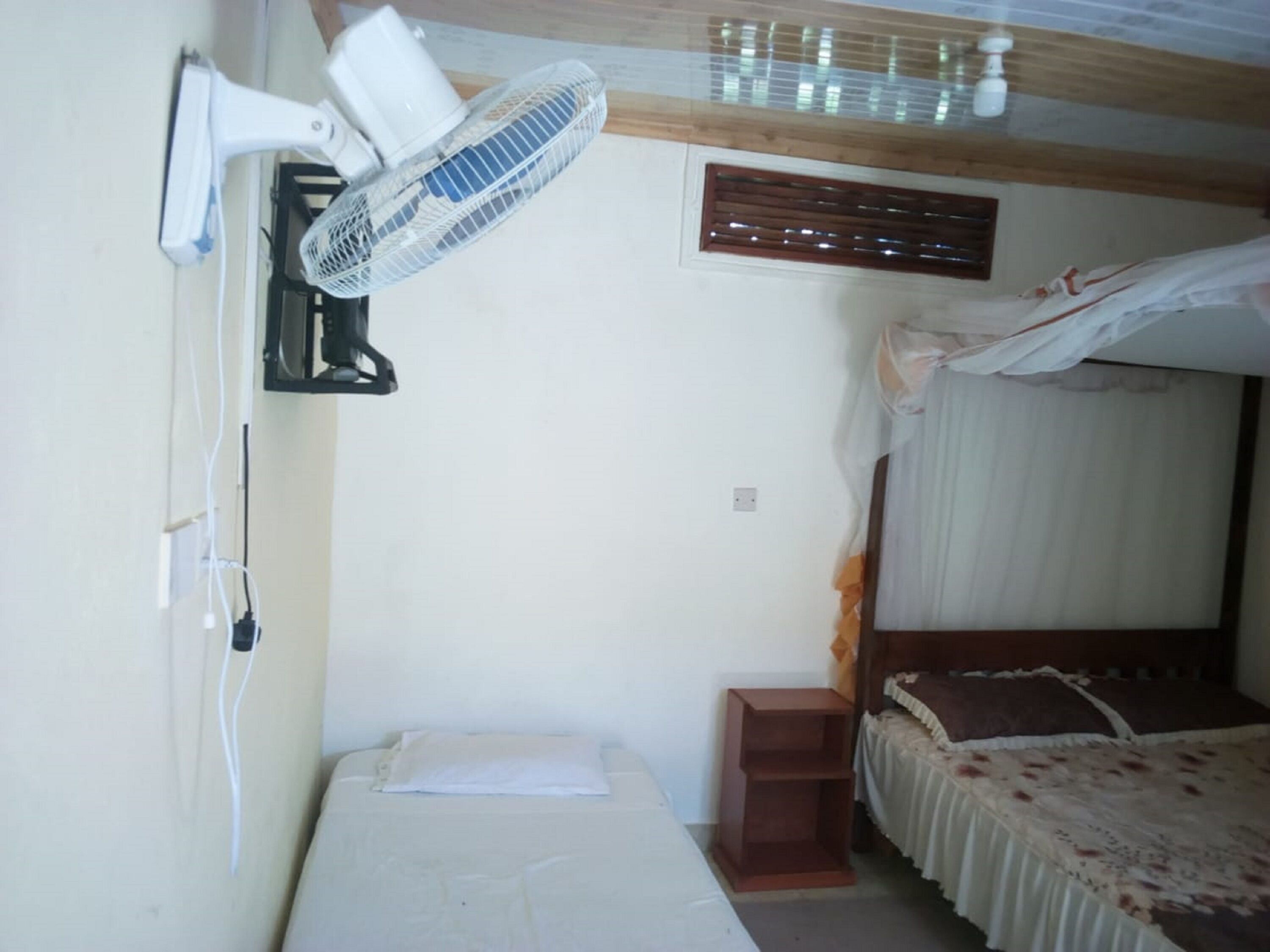 Een kamer met een bed, nachtkastje, een ventilator aan de muur en een plafondventilator.