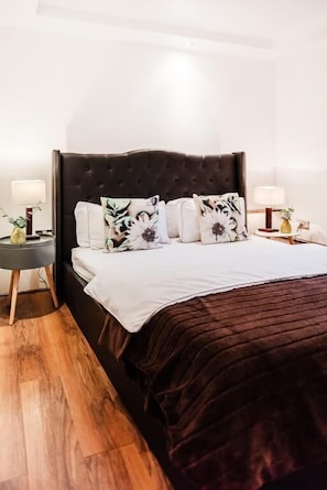 A neatly made bed with a dark headboard, white bedding, and floral-patterned pillows. There are two bedside tables with lamps and a vase on each. The room has wooden flooring and white walls. - My Lady (London)