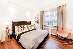 A bedroom with a large bed, a rocking chair, a window with curtains, and a bedside table with a lamp.