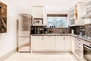A modern kitchen with white cabinetry, a stainless steel refrigerator, a microwave, and a window with a view of trees. - My Lady (London)