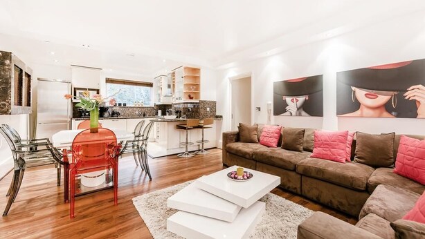A modern living room with a sofa, coffee table, and chairs. There is a kitchen in the background with a dining area. - My Lady (London)