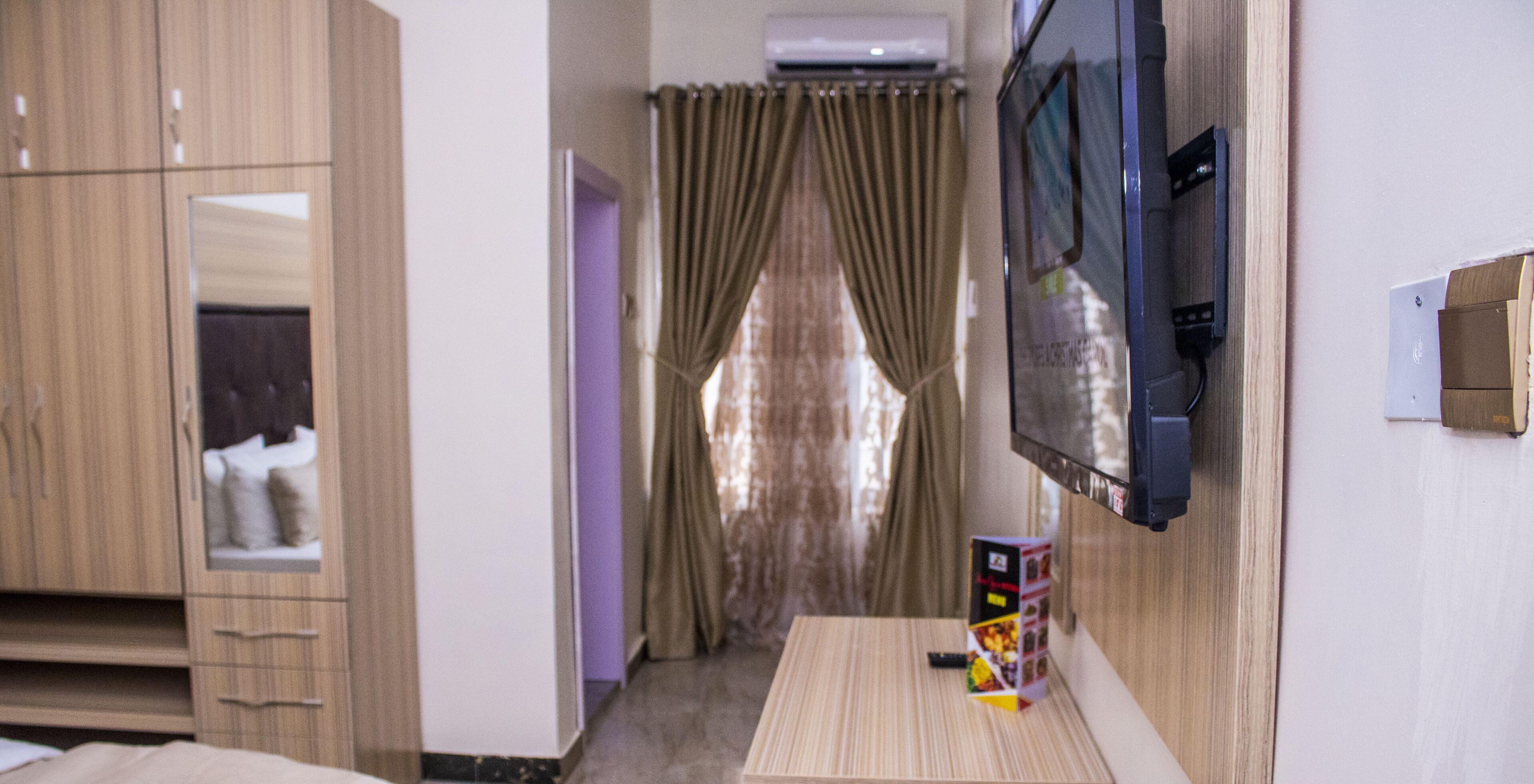 Habitación de hotel con un armario de madera, un televisor montado en la pared y una pequeña mesa con un control remoto y una revista.
