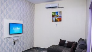 Een woonkamer met een wandgemonteerde flatscreen-tv waarop 'DStv London' wordt weergegeven, een luchtverwarmingssysteem aan de muur en een kleurrijk abstract schilderij.