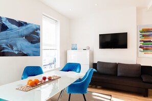 A modern living room with a white dining table, blue chairs, a black sofa, and a flat-screen TV mounted on the wall.