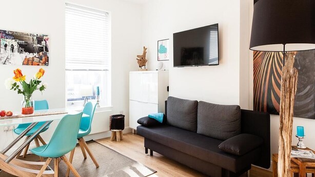 Una sala moderna con un sofá, un televisor montado en la pared, una mesa de comedor con sillas y una ventana con persianas.
