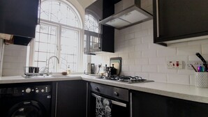 A kitchen with a dishwasher, stove, and a window with a view of trees. - Heritage Millworkers Cosy Cottage (Shipley)