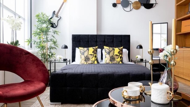 Une chambre moderne avec une tête de lit noire recouverte de tissu, une chaise rouge, une table de chevet avec une lampe et un élément décoratif mural.