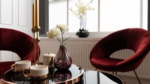 Une pièce avec une table ronde en verre, deux fauteuils en velours rouge, un plateau avec des tasses et un vase rempli de fleurs, ainsi qu’une fenêtre ornée de rideaux blancs et d’une plante.