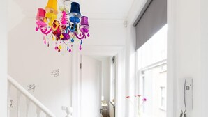 Un couloir lumineux, agrémenté d’un lustre coloré, de murs blancs et d’une fenêtre où sont disposées des plantes en pot.