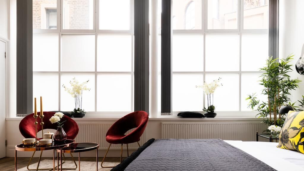 A modern hotel room with a large window, a bed, a red chair, a small table with a vase of flowers, and a potted plant.