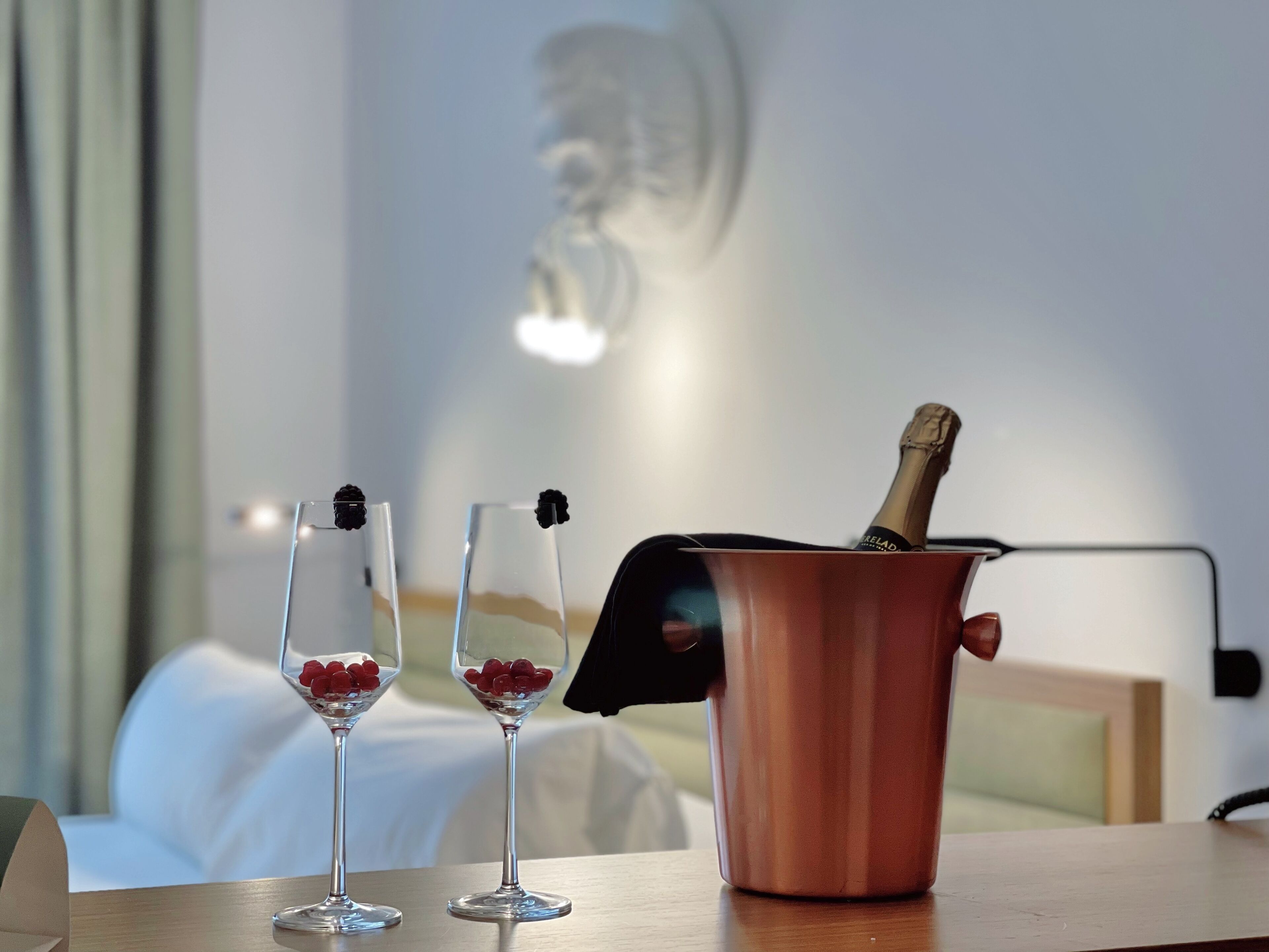 Dos copas de champán con frutos rojos sobre una mesa de madera, un balde de cobre con una botella de champán, y una cama al fondo.