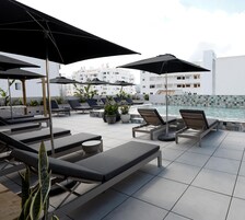 Un área de piscina en la azotea con sillones de descanso y sombrillas, piso de baldosas y vista a un edificio blanco al fondo.