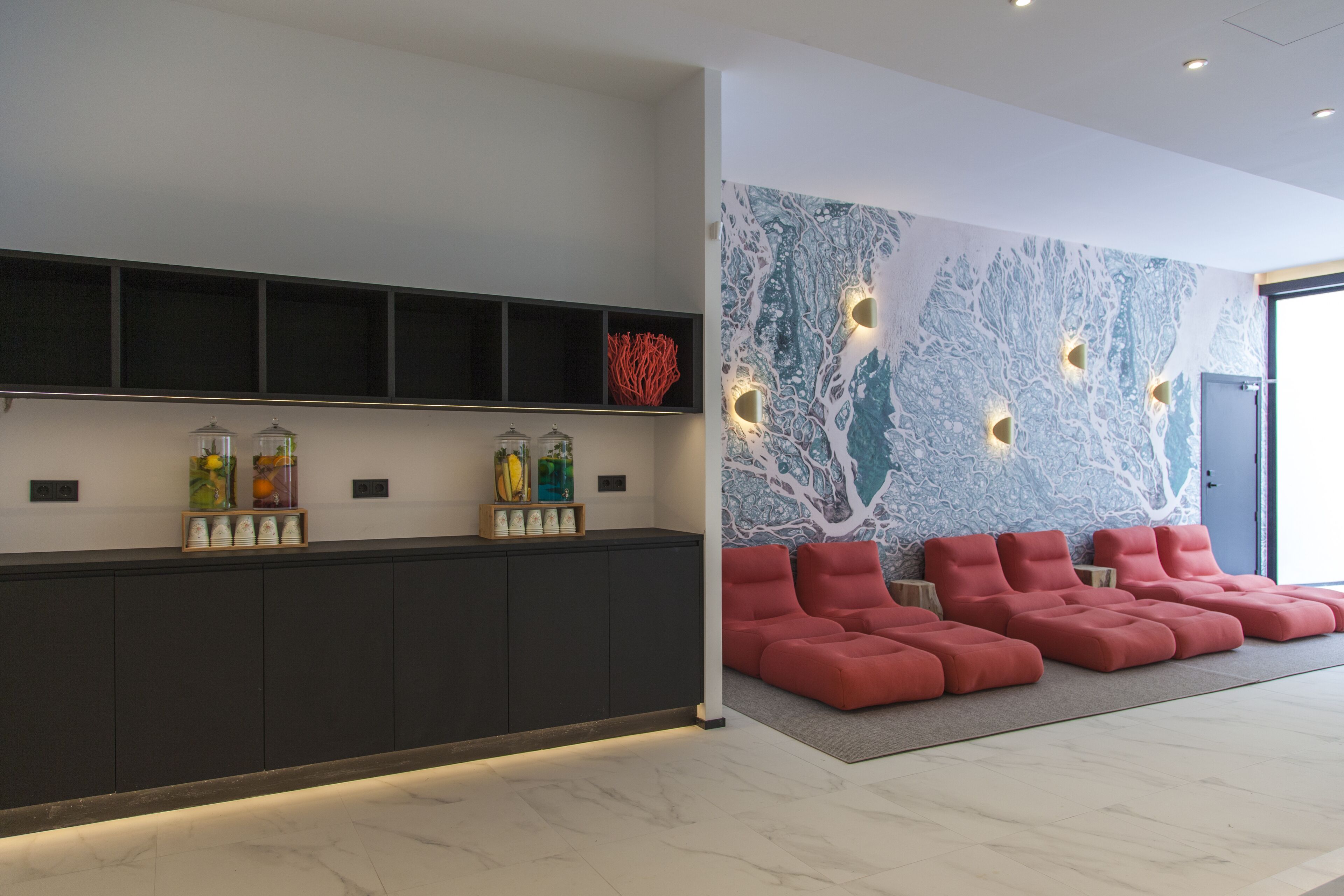 A reception area with a dark reception desk, shelves with decorative items, a wall with a tree mural, and red seating.