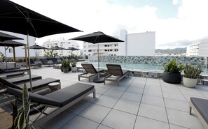 A rooftop pool area with lounge chairs and umbrellas, a tiled floor, and a view of buildings and mountains in the background. - Stic Urban Hotel & Spa (Sant Antoni de Portmany)