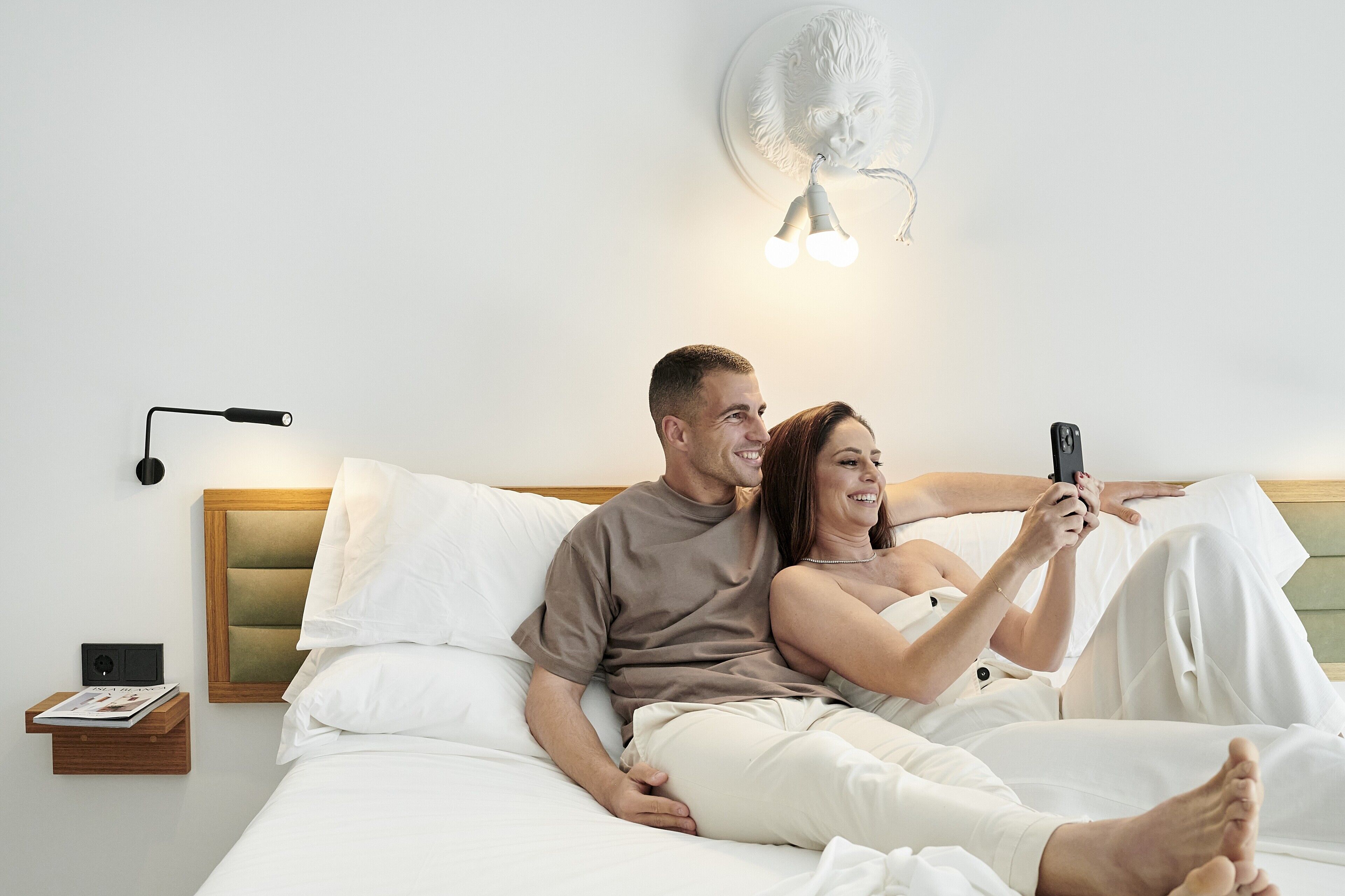 A couple in a bedroom with a bed, bedside table, and wall-mounted fan.