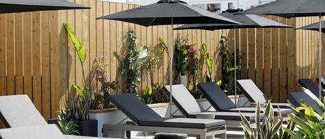 Un área de lounge en la azotea con sillones de sol y sombrillas, rodeada por vallas de madera y plantas en macetas.