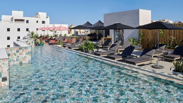 Un área de piscina en la azotea con sillones de descanso y sombrillas, rodeada de edificios.