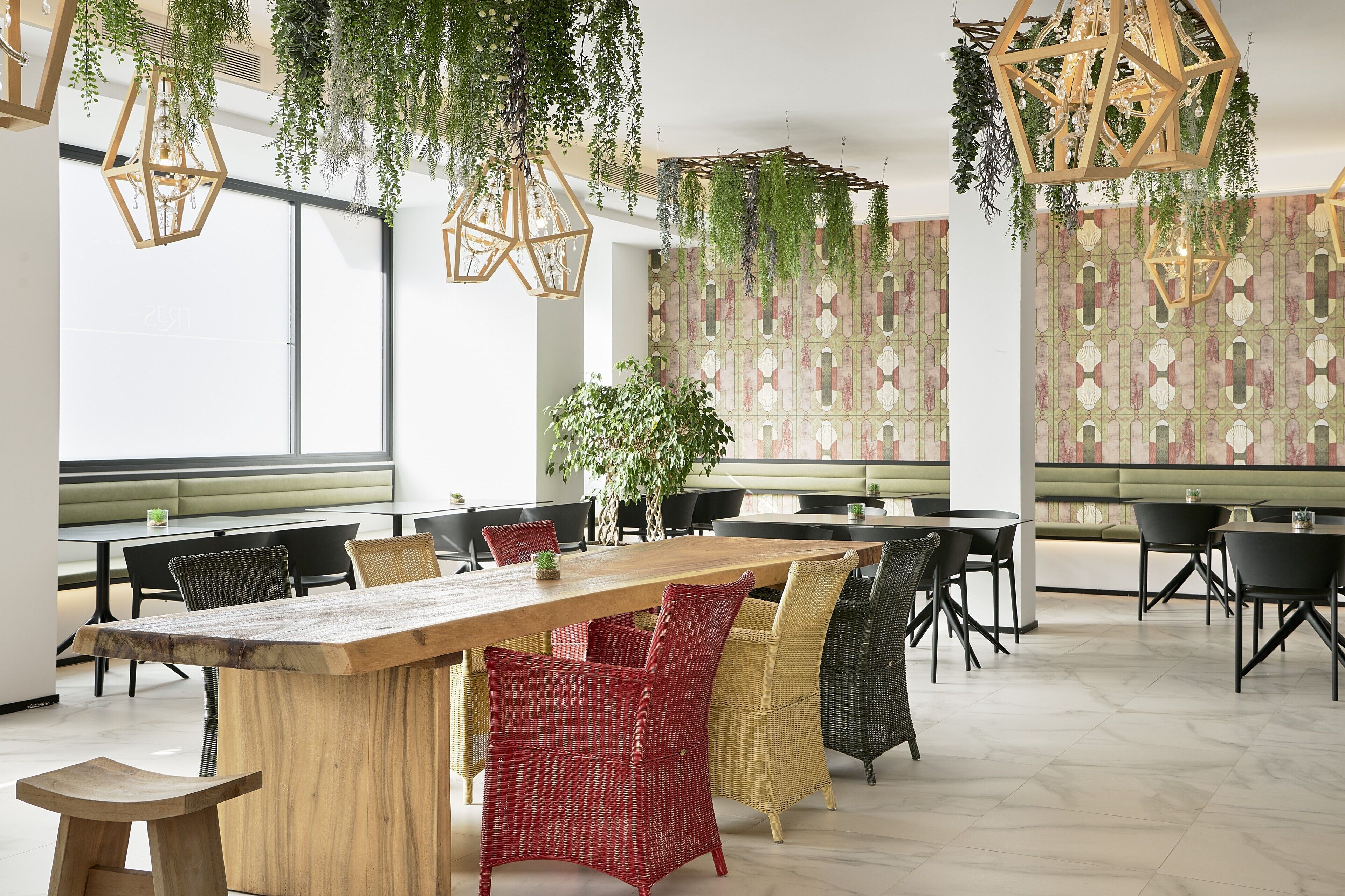 A modern dining area with a long wooden table, wicker chairs, hanging plants, and patterned wallpaper.
