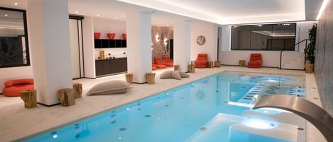 Une piscine intérieure moderne, avec une eau d’un bleu limpide, entourée de sièges rouges et agrémentée d’éléments décoratifs en bois.